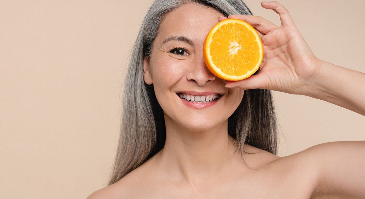 Das Bild zeigt eine lächelnde Frau mit grauem Haar, die eine Orangenhälfte spielerisch vor ihr rechtes Auge hält. Der Hintergrund ist ein einheitlicher beigefarbener Ton, der den Fokus auf die Person und die Frucht lenkt. Die Frau erscheint nackt zu sein. Ihre strahlende Haut und ihr fröhlicher Ausdruck vermitteln eine positive Assoziation mit dem Verzehr von frischem Obst und einer gesunden Ernährung.