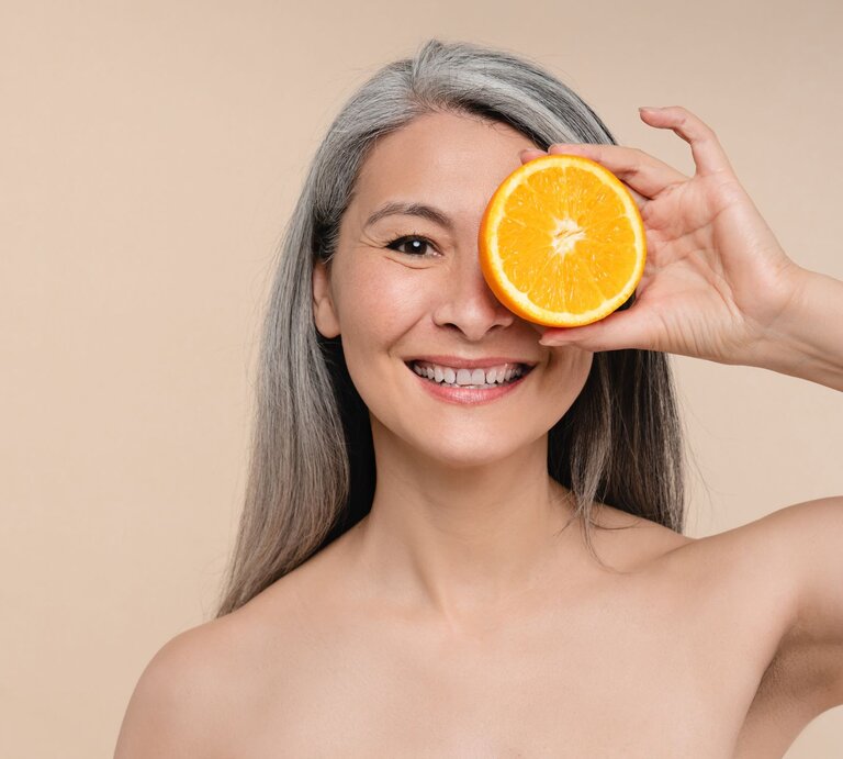 Das Bild zeigt eine lächelnde Frau mit grauem Haar, die eine Orangenhälfte spielerisch vor ihr rechtes Auge hält. Der Hintergrund ist ein einheitlicher beigefarbener Ton, der den Fokus auf die Person und die Frucht lenkt. Die Frau erscheint nackt zu sein. Ihre strahlende Haut und ihr fröhlicher Ausdruck vermitteln eine positive Assoziation mit dem Verzehr von frischem Obst und einer gesunden Ernährung. | © Adobe Stock/ InsideCreativeHouse