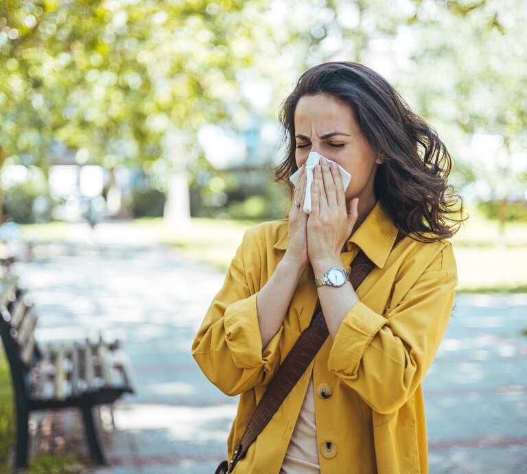 Eine Frau mit dunklen Haaren und gelbem Mantel niest aufgrund von Heuschnupfen  mit geschlossenen Augen in ein Taschentuch in einem sonnigen Park. | © Shutterstock/Dragana Gordic