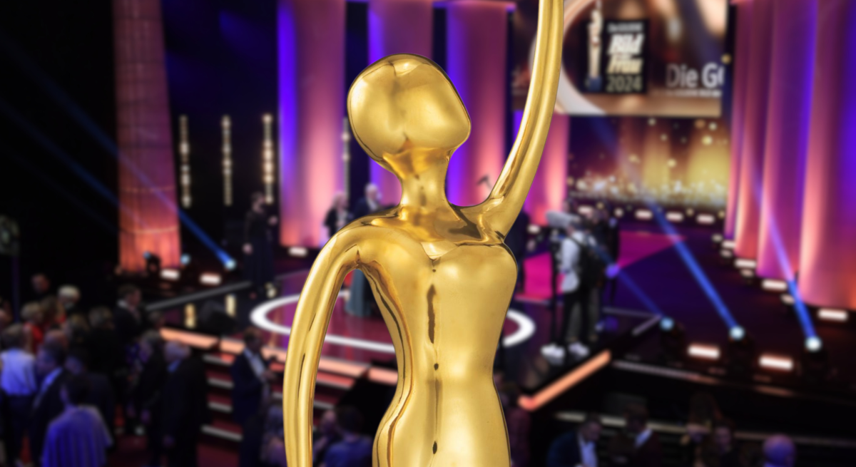 GOLDENE BILD der FRAU Statue mit erhobenem Arm vor beleuchteter Bühne und Publikum bei festlicher Gala mit lila Lichtakzenten.