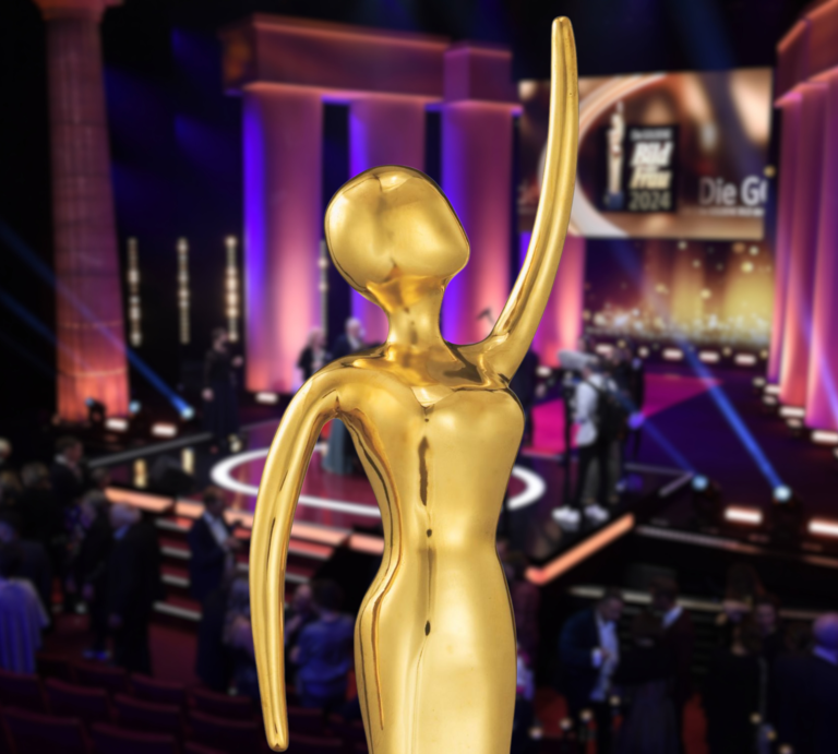 GOLDENE BILD der FRAU Statue mit erhobenem Arm vor beleuchteter Bühne und Publikum bei festlicher Gala mit lila Lichtakzenten. | ©  IMAGO / Marja; Goldene BILD der FRA; Collage: BILD der FRAU