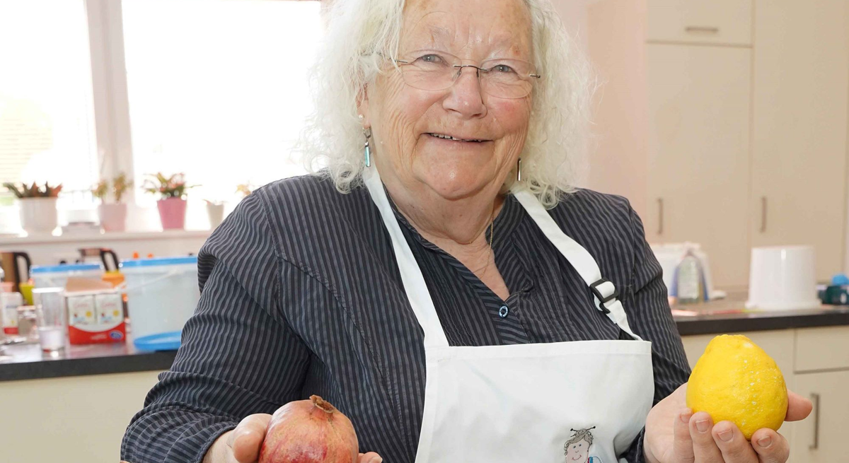 Ältere Frau mit weißen Locken und Brille lächelt in Küche, hält Granatapfel und Zitrone, trägt gestreifte Bluse und Schürze.