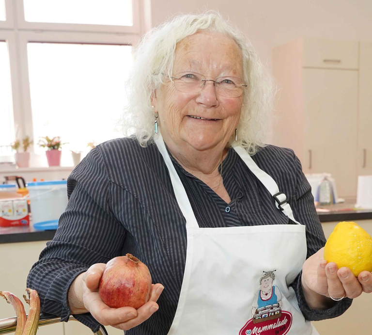 Ältere Frau mit weißen Locken und Brille lächelt in Küche, hält Granatapfel und Zitrone, trägt gestreifte Bluse und Schürze. | © GOLDENE BILD der FRAU