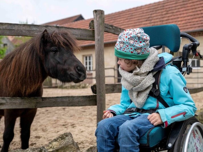 Kind im Rollstuhl mit bunter Mütze und Winterjacke blickt neugierig ein schwarzes Pony an, das hinter einem Holzzaun steht. | © GOLDENE BILD der FRAU