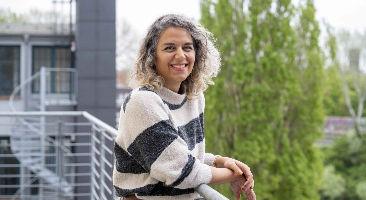 Junge Frau mit grauen Locken und gestreiftem Pullover lehnt lächelnd am Balkongeländer vor grünen Bäumen und moderner Architektur.