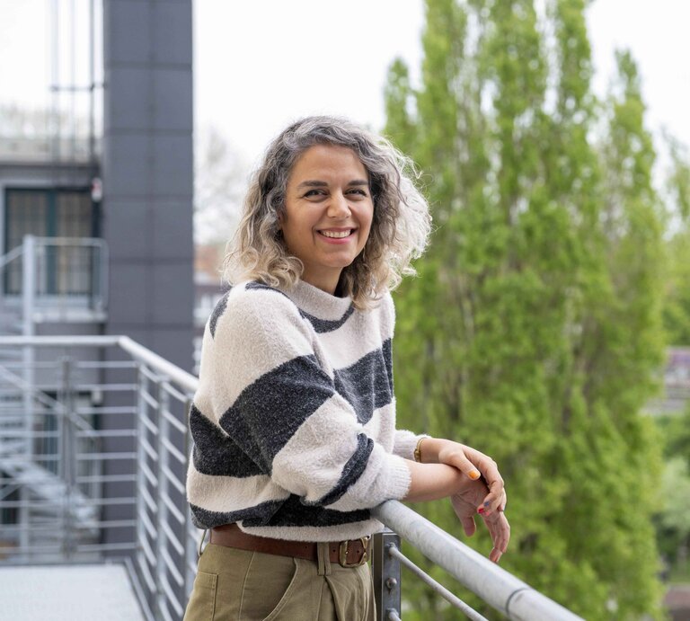 Junge Frau mit grauen Locken und gestreiftem Pullover lehnt lächelnd am Balkongeländer vor grünen Bäumen und moderner Architektur. | © GOLDENE BILD der FRAU