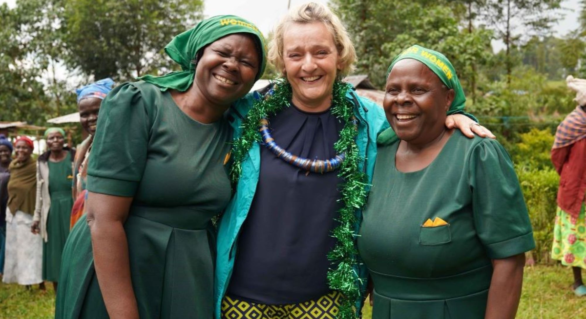 Glückliche Szene: Drei Frauen umarmen sich im Grünen, lachen in die Kamera, im Hintergrund weitere Frauen in traditioneller Kleidung.