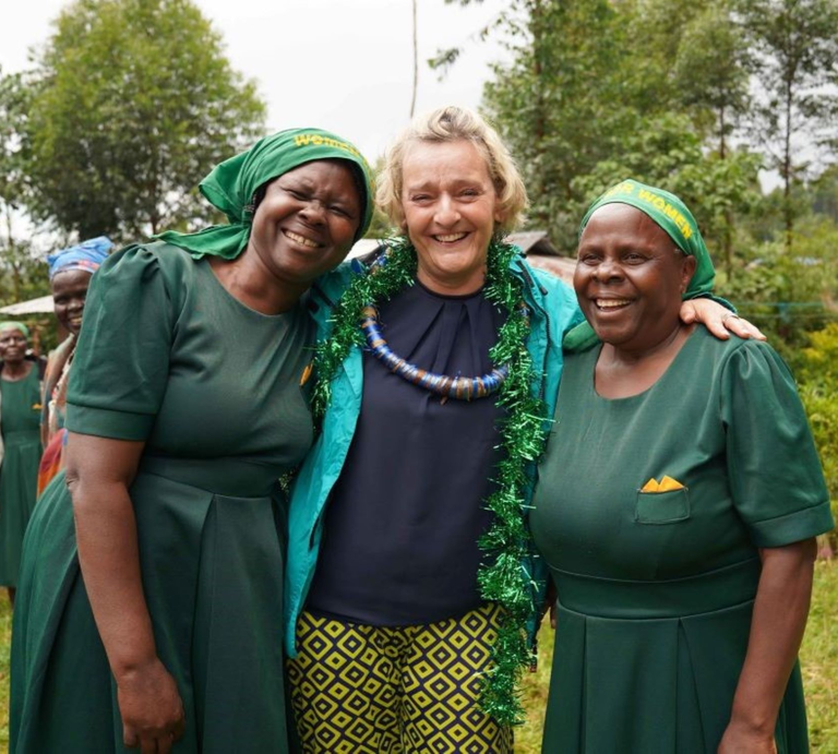 Glückliche Szene: Drei Frauen umarmen sich im Grünen, lachen in die Kamera, im Hintergrund weitere Frauen in traditioneller Kleidung. | © GOLDENE BILD der FRAU