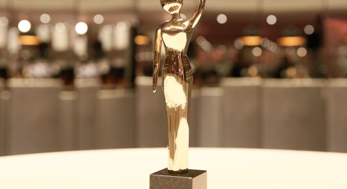 Eine glänzende, goldene Trophäe in menschlicher Form steht auf einem schwarzen Sockel vor einem verschwommenen, hell erleuchteten Hintergrund.