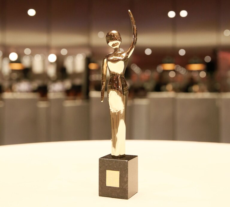 Eine glänzende, goldene Trophäe in menschlicher Form steht auf einem schwarzen Sockel vor einem verschwommenen, hell erleuchteten Hintergrund. | © GOLDENE BILD der FRAU