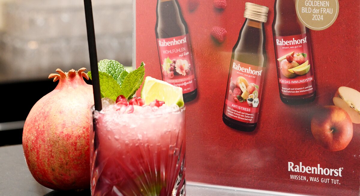 Ein fruchtiger Cocktail mit Granatapfelkernen, Minze und Limette steht vor einem roten Werbeaufsteller von Rabenhorst auf einer dunklen Theke.
