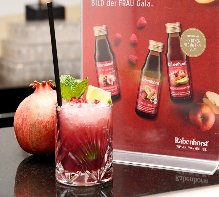Ein fruchtiger Cocktail mit Granatapfelkernen, Minze und Limette steht vor einem roten Werbeaufsteller von Rabenhorst auf einer dunklen Theke. | © Karin Costanzo/BILD der FRAU