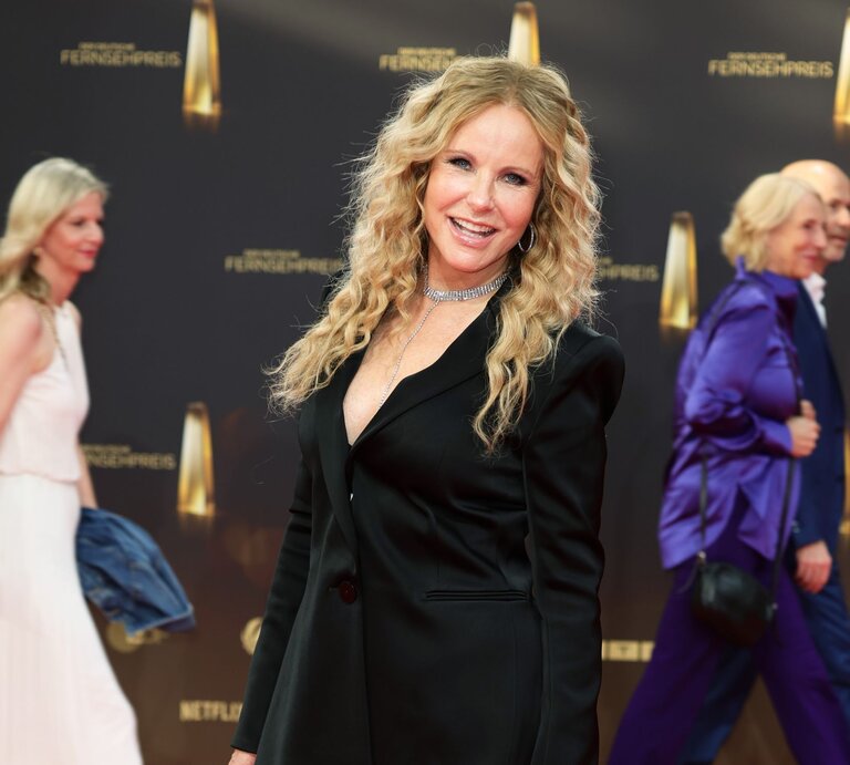 Blonde Frau mit langen, lockigen Haaren und schwarzem Blazer lächelt auf dem roten Teppich, dahinter laufen zwei elegant gekleidete Personen vorbei. | © Getty Images