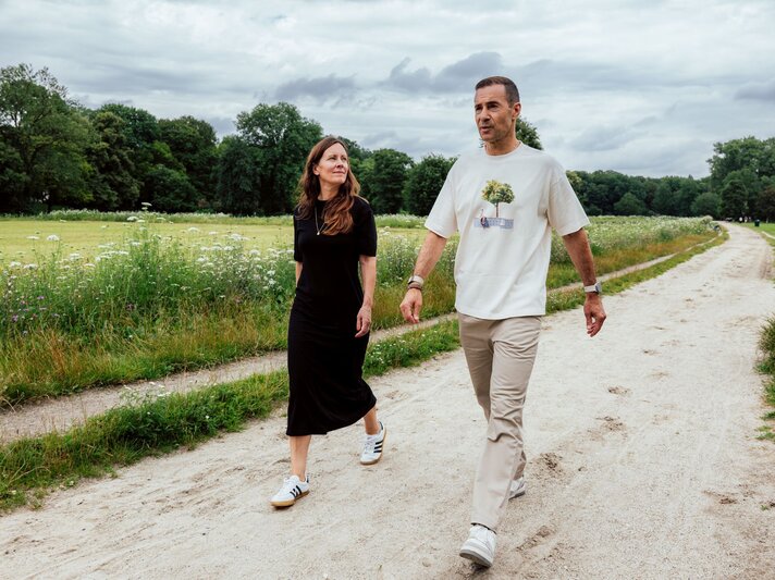 Eine Frau in schwarzem Kleid und ein Mann in hellem Shirt spazieren bei bewölktem Himmel auf einem Sandweg durch einen grünen Park. | © Maximilian Ralf Schmidt