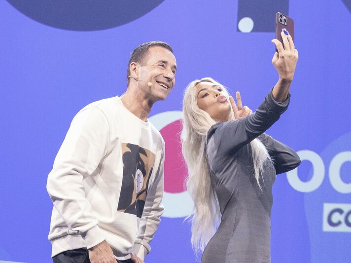 Ein Mann in weißem Pullover und eine blonde Frau in grauem Kleid machen lachend ein Selfie auf einer Bühne. | © dpa