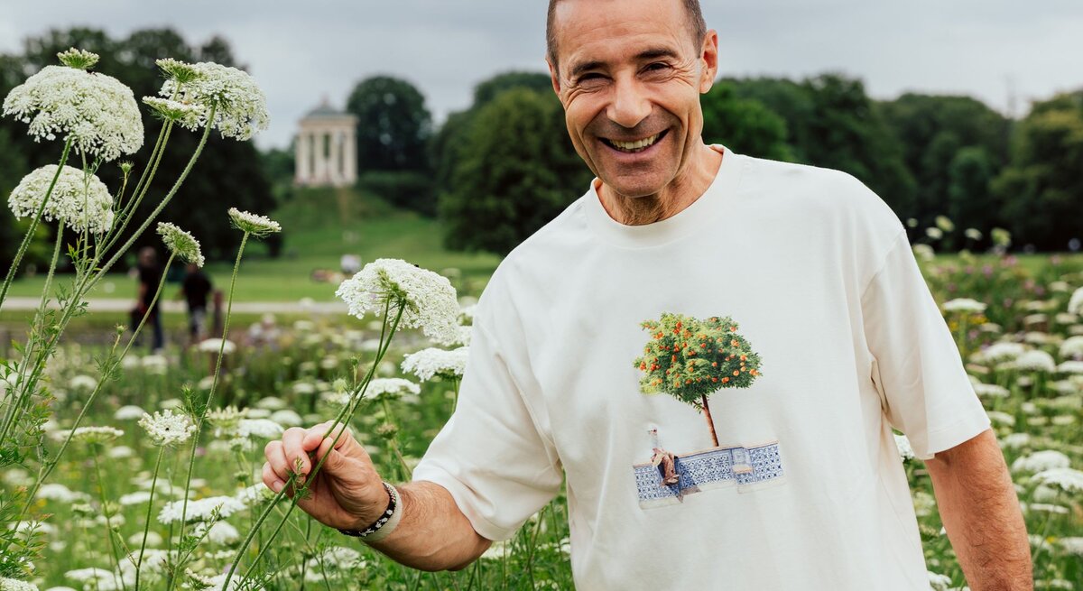 Ein lächelnder Mann mittleren Alters mit kurzen braunen Haaren steht in einer Wiese mit weißen Blüten vor einem bewölkten Himmel.