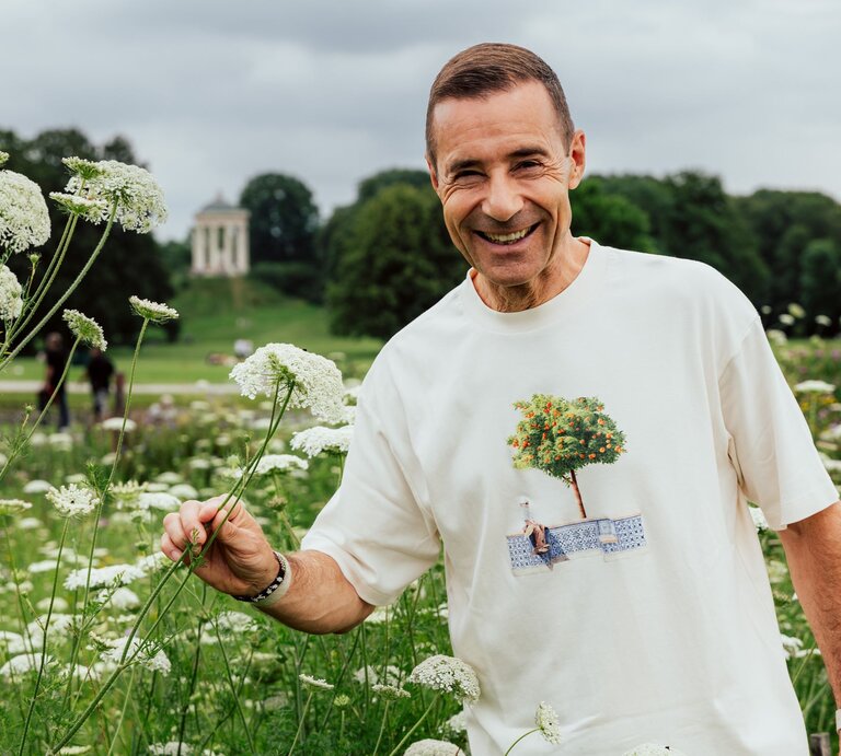 Ein lächelnder Mann mittleren Alters mit kurzen braunen Haaren steht in einer Wiese mit weißen Blüten vor einem bewölkten Himmel. | © Maximilian Ralf Schmidt