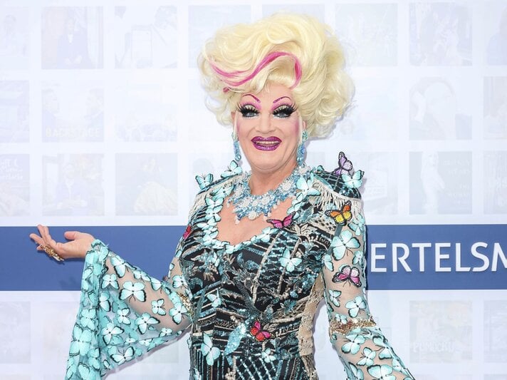 Bunt geschminkte Drag-Queen mit großer, blonder Perücke und farbenfrohem Kleid voller Schmetterlinge lächelt. | © dpa