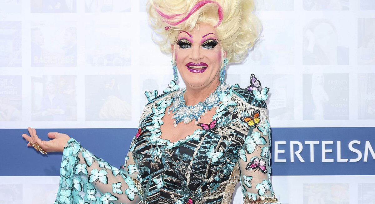 Bunt geschminkte Drag-Queen mit großer, blonder Perücke und farbenfrohem Kleid voller Schmetterlinge lächelt.