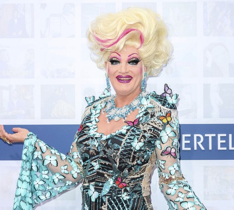 Bunt geschminkte Drag-Queen mit großer, blonder Perücke und farbenfrohem Kleid voller Schmetterlinge lächelt. | © dpa