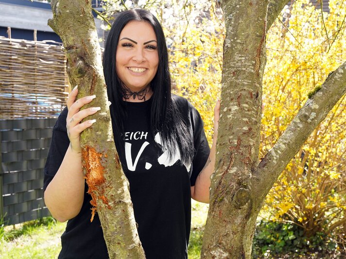 Frau mit langen schwarzen Haaren, tätowiertem Hals und schwarzem Shirt lächelt zwischen zwei Bäumen im Garten. | © Marion Vogel