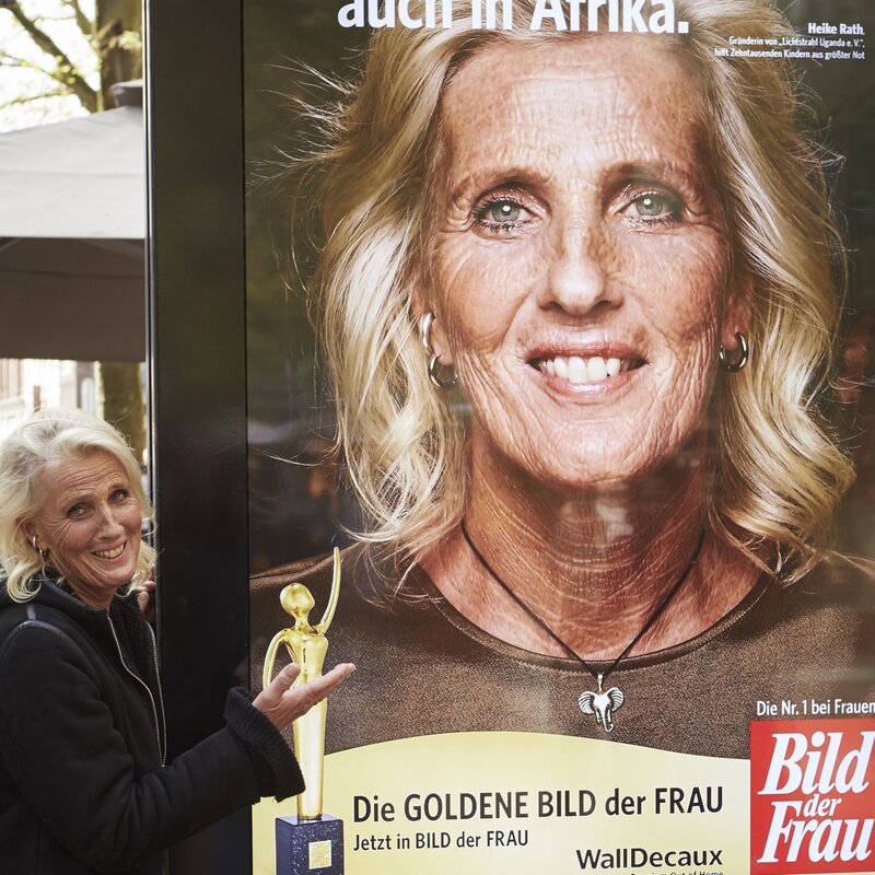 Eine ältere Frau mit schulterlangem blondem Haar und schwarzer Jacke präsentiert lächelnd ein Werbeplakat der "Goldene Bild der Frau" auf einem belebten Platz. | © dsphotos.de