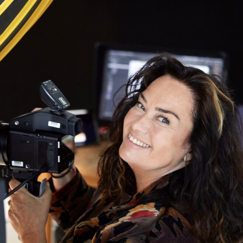Lachende Frau mit dunklen, welligen Haaren und einem Camouflage-Mantel hält eine Kamera in einem Studio mit professioneller Beleuchtung. | © dsphotos.de