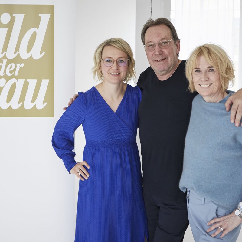 Drei lächelnde Personen stehen in einem hellen Raum vor einem "Bild der Frau"-Banner, zwei Frauen und ein Mann, Arm in Arm. | © dsphotos.de