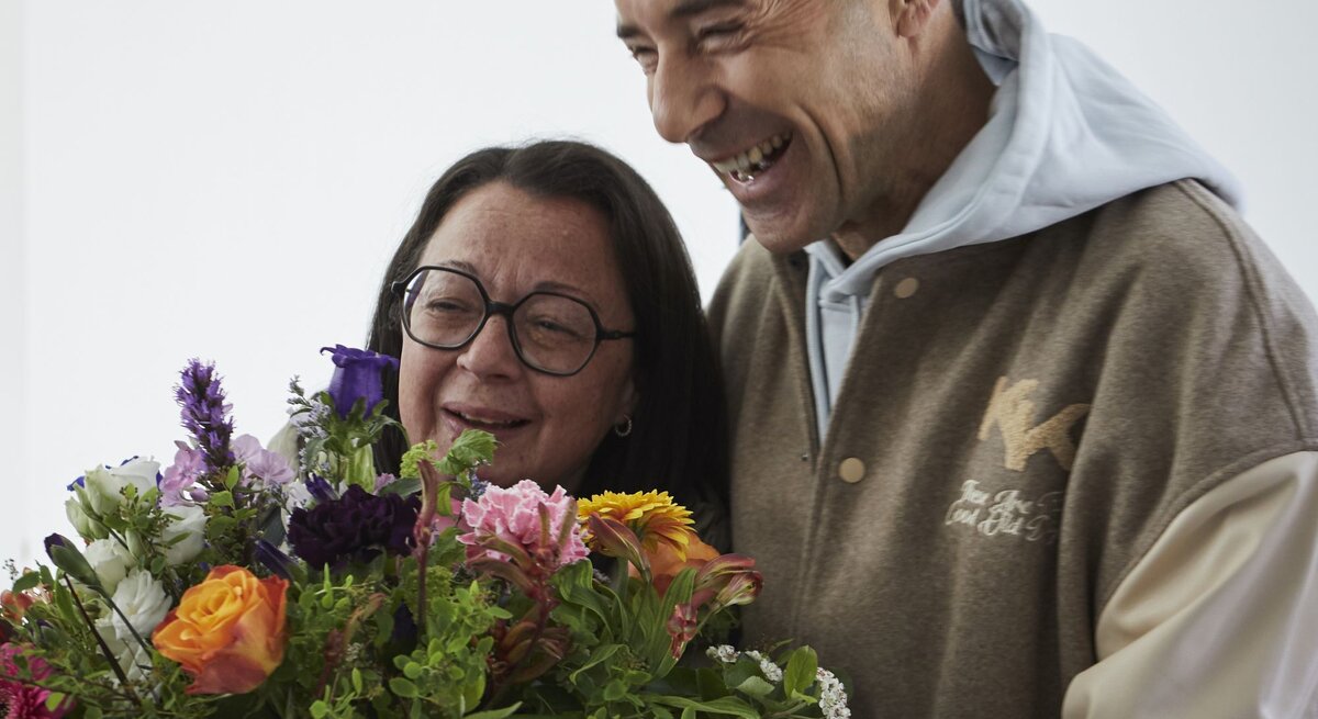Eine ältere Frau mit Brille und ein lachender Mann in beiger Jacke halten gemeinsam einen bunten Blumenstrauß.