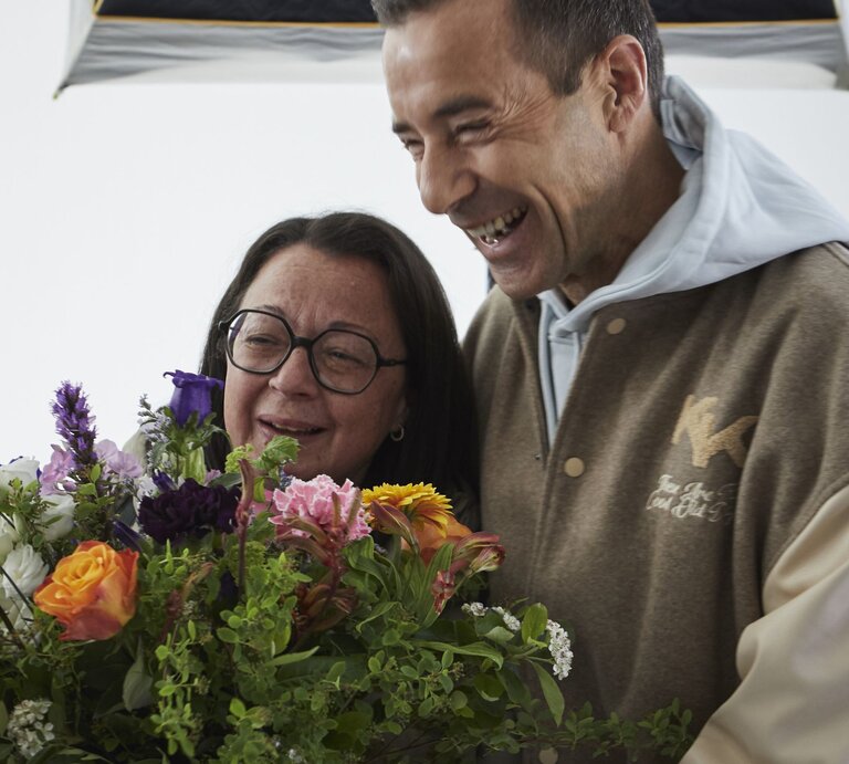 Eine ältere Frau mit Brille und ein lachender Mann in beiger Jacke halten gemeinsam einen bunten Blumenstrauß. | © dsphotos.de