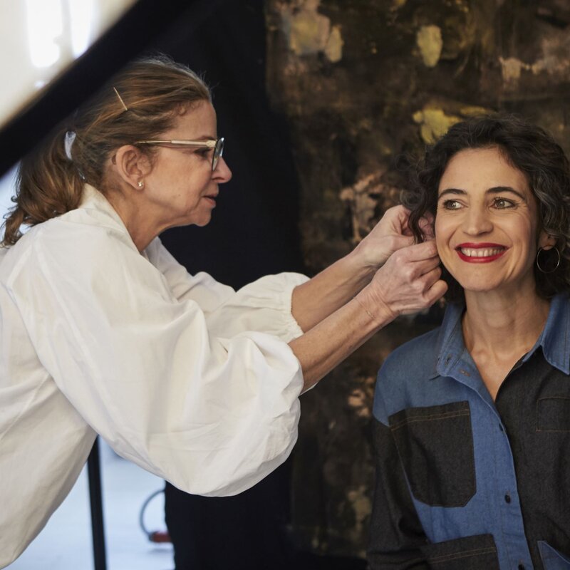 Visagistin mit Brille und weißer Bluse richtet Haare einer lächelnden Frau mit lockigem Haar und rotem Lippenstift in einem Studio. | © dsphotos.de