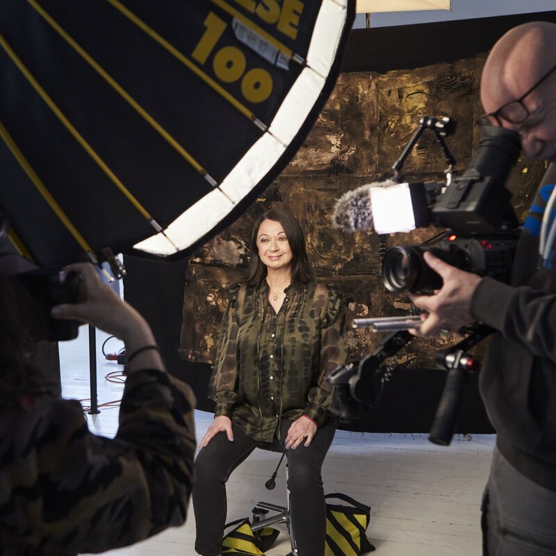 Eine Frau mittleren Alters in grünem Camouflagehemd sitzt lächelnd vor einer Kamera, während zwei Personen sie fotografieren und filmen. | © dsphotos.de