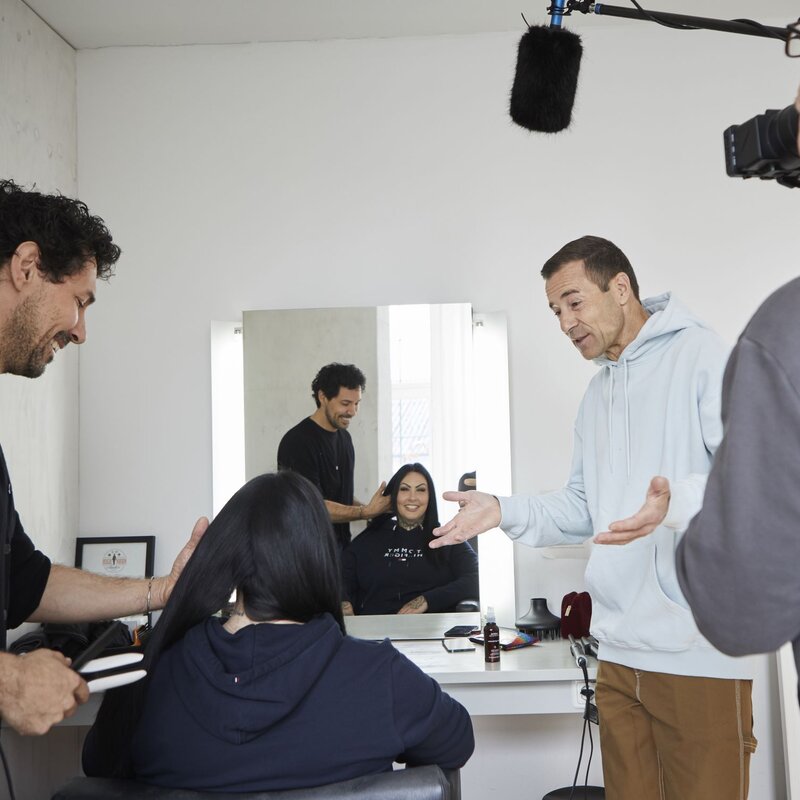 Visagist und Mann im hellen Hoodie unterhalten sich, während sie die Haare einer Frau im Friseursalon stylen und ein Kameramann aufnimmt. | © dsphotos.de