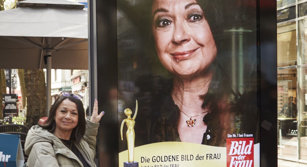 Eine ältere Frau in beiger Jacke präsentiert lächelnd ein großes Werbeplakat der "Goldene Bild der Frau" in einer belebten Fußgängerzone.