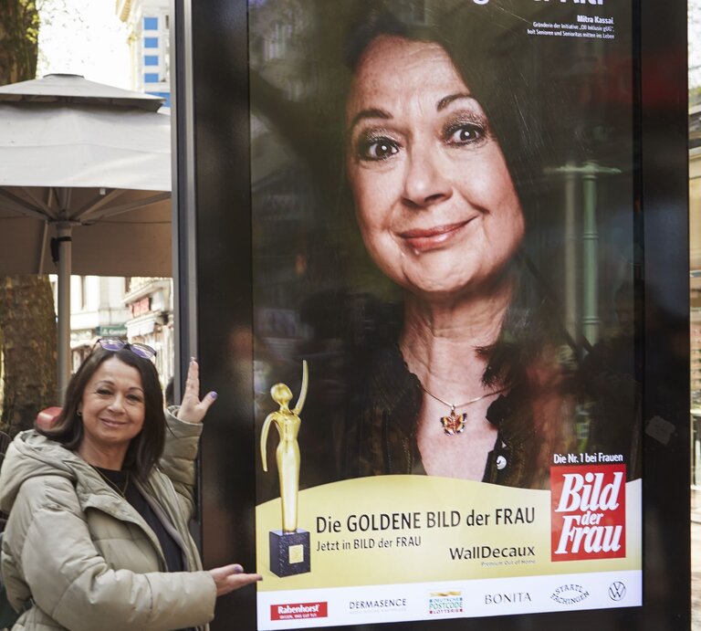Eine ältere Frau in beiger Jacke präsentiert lächelnd ein großes Werbeplakat der "Goldene Bild der Frau" in einer belebten Fußgängerzone. | © dsphotos.de