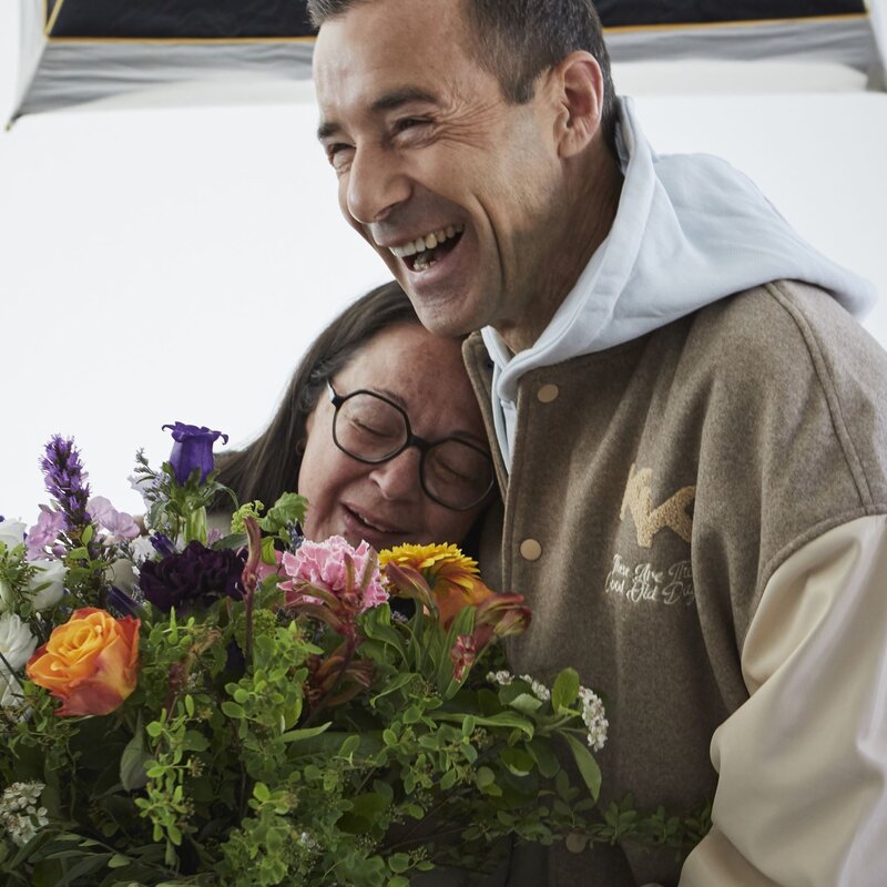Ein lachender Mann in beiger Jacke umarmt eine ältere Frau mit Brille und dunklen Haaren, die einen bunten Blumenstrauß hält. | © dsphotos.de