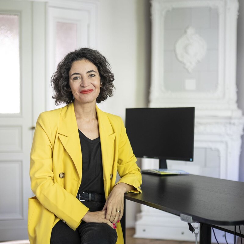 Eine Frau mittleren Alters mit lockigem dunklem Haar und rotem Lippenstift lächelt in gelbem Blazer in einem hellen Büro. | © dsphotos.de
