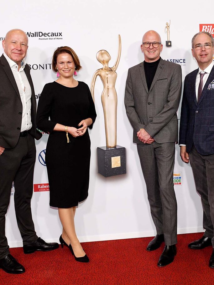 Drei Männer in Anzügen und eine Frau in schwarzem Kleid stehen lächelnd auf rotem Teppich neben einer goldenen Trophäe vor einer Sponsorenwand. | © BILD der FRAU 