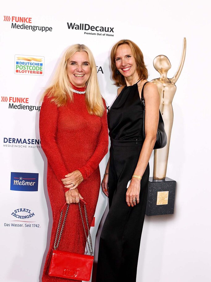 Zwei Frauen in eleganten Abendkleidern, eine in Rot mit Glitzerstiefeln, lächeln auf rotem Teppich vor einer goldenen Statue und Sponsorenwand. | © BILD der FRAU 