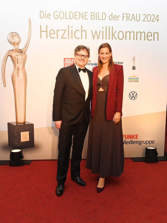 Elegant gekleidetes Duo posiert auf einem Gala-Event vor einer goldenen Trophäe und einer Wand mit dem Schriftzug "Goldene Bild der Frau 2024" | © BILD der FRAU 