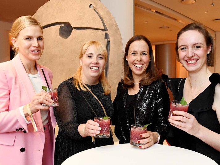 Vier fröhliche Frauen in festlicher Kleidung halten Cocktails in der Hand und stehen lachend an einem weißen Stehtisch in modernem Ambiente. | © BILD der FRAU 
