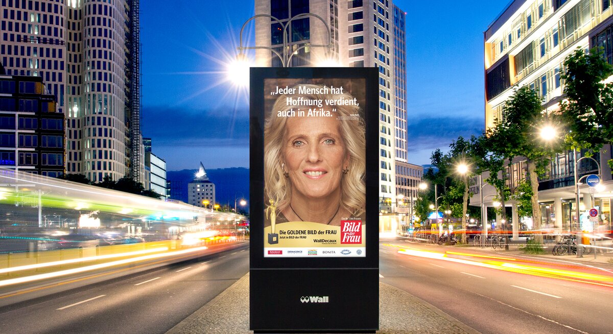 Leuchtendes Werbeplakat mit dem Bild einer blonden Frau und einem Hoffnungszitat, umgeben von modernen Hochhäusern in der Dämmerung.