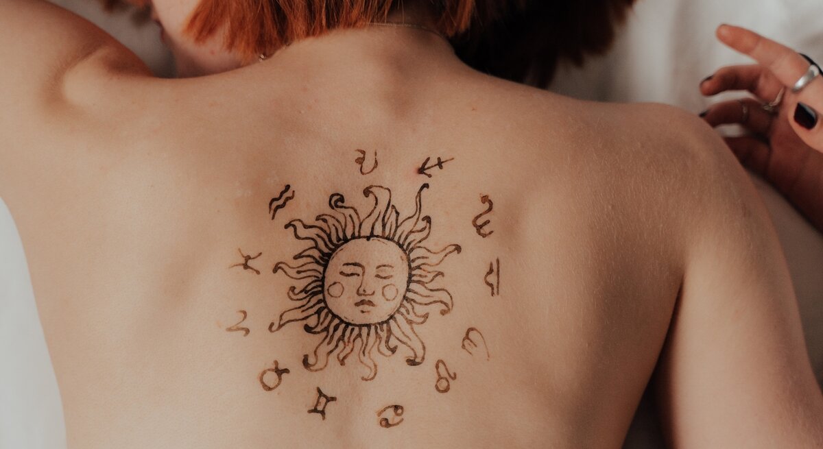 Eine Person mit roten Haaren liegt auf dem Bauch, während ihr Rücken ein temporäres oder permanentes Tattoo mit einer Sonne und astrologischen Symbolen ziert. Die Sonne im Zentrum des Designs hat ein Gesicht und wird von den zwölf Tierkreiszeichen umgeben. Die Person trägt Schmuck an der Hand und befindet sich in einer entspannten Haltung.