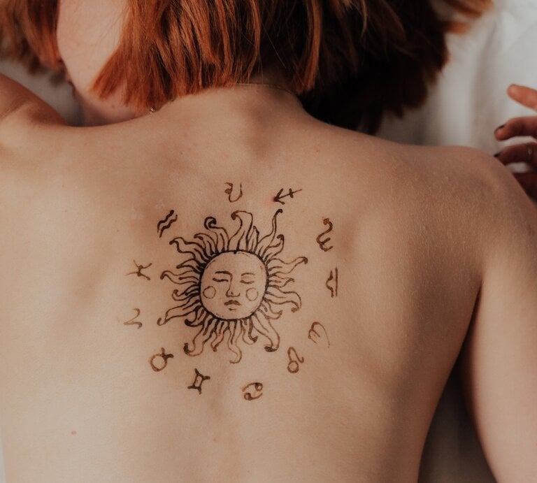 Eine Person mit roten Haaren liegt auf dem Bauch, während ihr Rücken ein temporäres oder permanentes Tattoo mit einer Sonne und astrologischen Symbolen ziert. Die Sonne im Zentrum des Designs hat ein Gesicht und wird von den zwölf Tierkreiszeichen umgeben. Die Person trägt Schmuck an der Hand und befindet sich in einer entspannten Haltung. | © Luca Paul Droß von capturenow via Canva.com