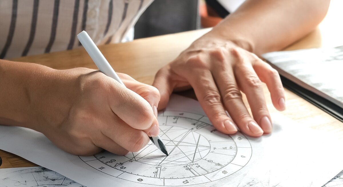 Eine Person berechnet ein Horoskop per Hand, indem sie mit einem Stift auf einem astrologischen Geburtshoroskop zeichnet. Die detaillierte Kreisgrafik enthält verschiedene astrologische Symbole und Linien, die Sternenkonstellationen darstellen. Der Arbeitsplatz ist mit weiteren astrologischen Unterlagen ausgestattet.