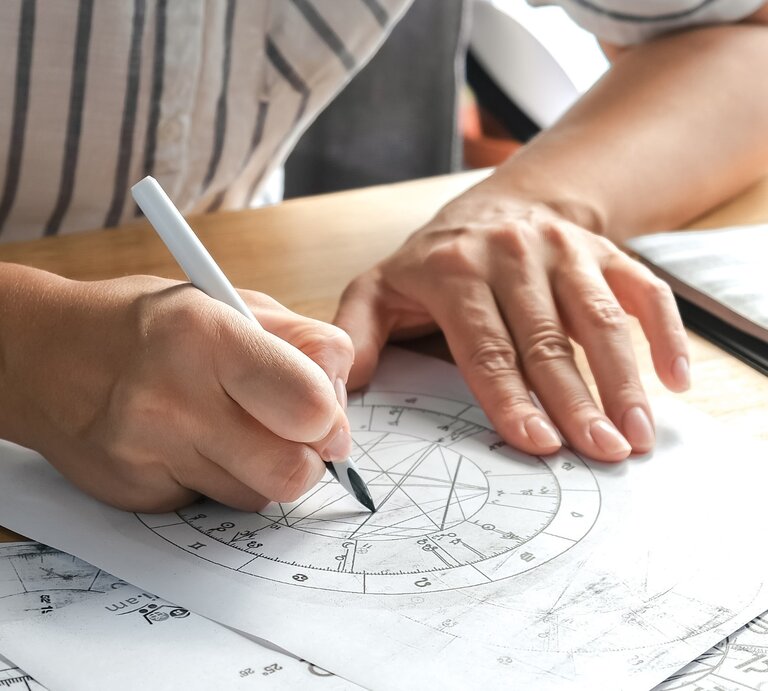 Eine Person berechnet ein Horoskop per Hand, indem sie mit einem Stift auf einem astrologischen Geburtshoroskop zeichnet. Die detaillierte Kreisgrafik enthält verschiedene astrologische Symbole und Linien, die Sternenkonstellationen darstellen. Der Arbeitsplatz ist mit weiteren astrologischen Unterlagen ausgestattet. | © Getty Images/ Shintartanya via Canva.com 