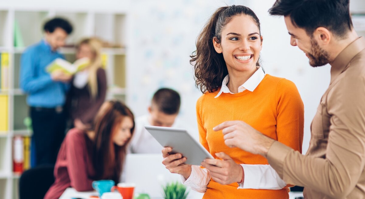 Eine junge Frau mit einem orangefarbenen Pullover und weißem Hemd hält ein Tablet und lächelt freundlich, während sie mit einem Kollegen im braunen Hemd spricht. Sie befinden sich in einem modernen, offenen Büro mit weiteren Mitarbeitern im Hintergrund, die sich austauschen oder konzentriert arbeiten. Die Atmosphäre wirkt dynamisch, kommunikativ und harmonisch.