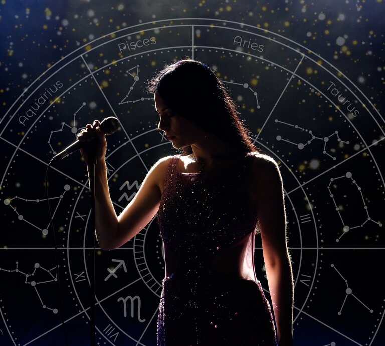 Eine Sängerin steht auf einer dunklen Bühne und hält ein Mikrofon in der Hand. Sie trägt ein funkelndes, elegantes Kleid, und ihr Gesicht ist teilweise im Schatten. Im Hintergrund leuchtet ein astrologisches Sternzeichen-Rad mit Konstellationen und Tierkreisnamen in goldenen Akzenten. Die Szene wirkt mystisch und symbolisiert eine Verbindung zwischen Kreativität, Talent und astrologischem Einfluss. | © Getty Image/ Nagaiets, WinWin.artlab und Mas Water via Canva.com [M]