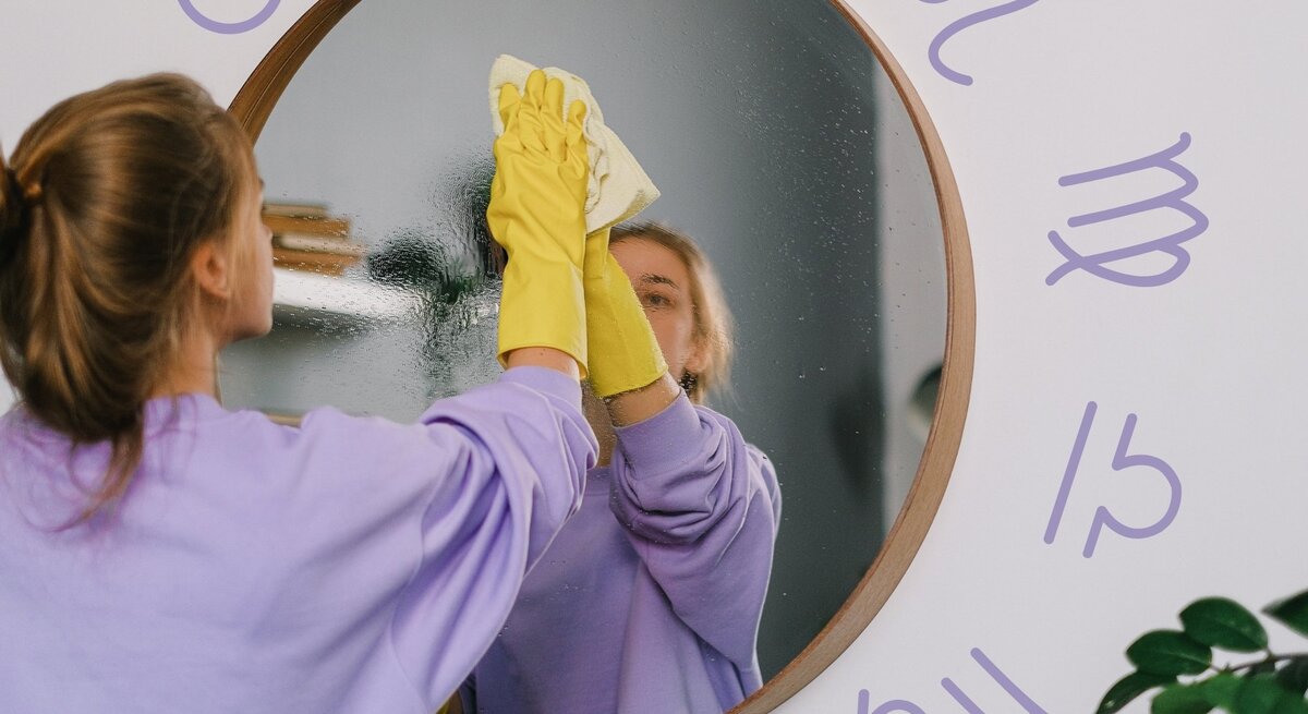 Eine junge Frau mit braunen Haaren, die zu einem lockeren Zopf gebunden sind, trägt einen lila Pullover und gelbe Gummihandschuhe, während sie einen runden Spiegel mit einem Tuch reinigt. Ihr Spiegelbild ist gut sichtbar. Der Hintergrund ist eine helle Wand, auf der astrologische Sternzeichen in lila gezeichnet sind. Die Szene vermittelt ein Gefühl von Ordnung und Reinigung, sowohl im physischen als auch im symbolischen Sinne.