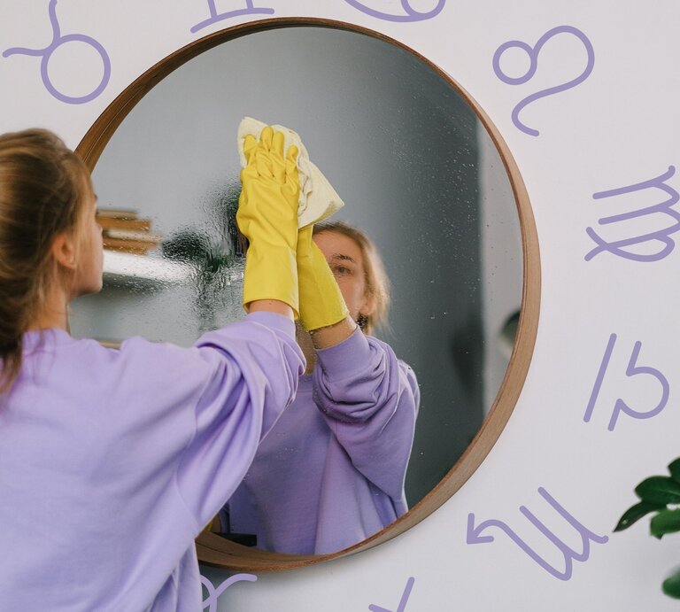 Eine junge Frau mit braunen Haaren, die zu einem lockeren Zopf gebunden sind, trägt einen lila Pullover und gelbe Gummihandschuhe, während sie einen runden Spiegel mit einem Tuch reinigt. Ihr Spiegelbild ist gut sichtbar. Der Hintergrund ist eine helle Wand, auf der astrologische Sternzeichen in lila gezeichnet sind. Die Szene vermittelt ein Gefühl von Ordnung und Reinigung, sowohl im physischen als auch im symbolischen Sinne. | © Pexels/ SHVETS production und Keronyart's Images via Canva.com [M]